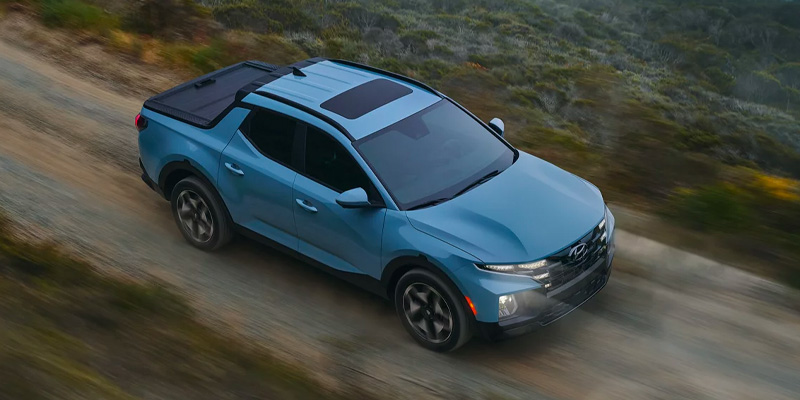 Blue Hyundai Santa Cruz pickup truck driving on a dirt road from an aerial view.